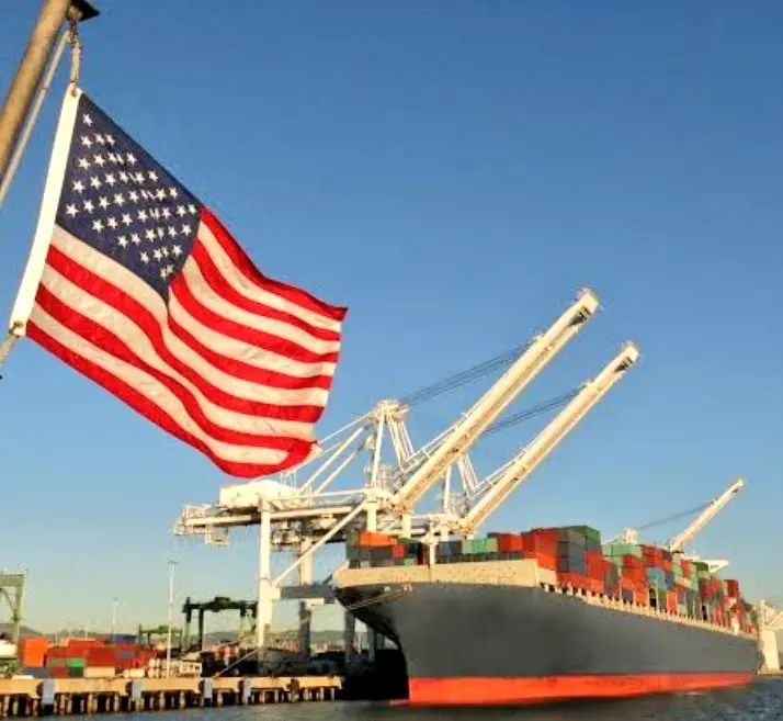U.S. flag flying beside a container ship being loaded at a port, illustrating new U.S. port-fee policies.