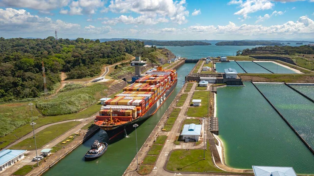 Large container ship passing through a canal lock, with a tugboat nearby and lush green surroundings on a sunny day