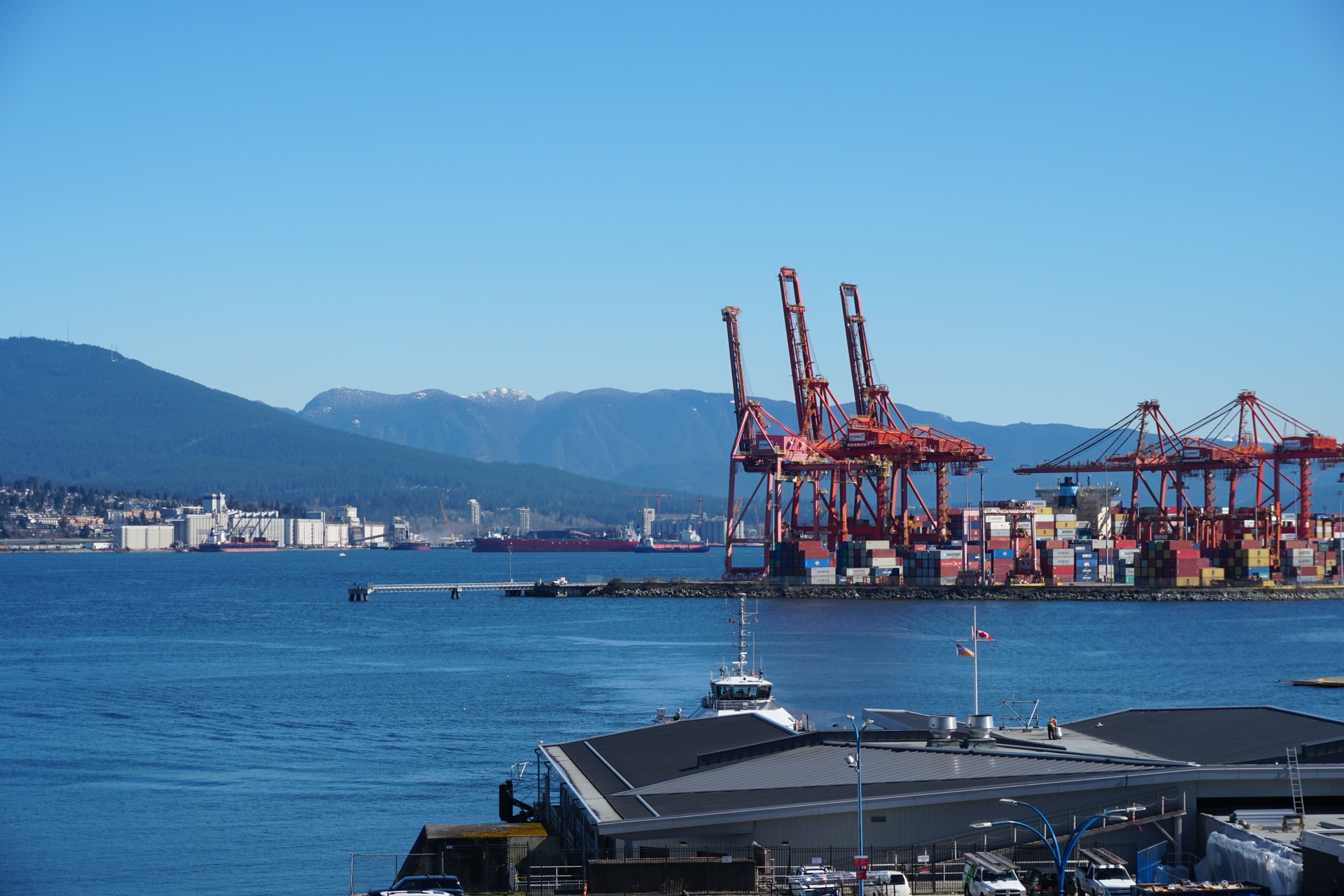Port scene with red gantry cranes and stacked cargo containers along the waterfront, mountains in the distance under a clear blue sky.
